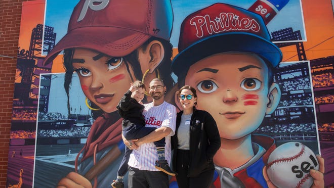 Colombia-born South Jersey artist Jose Bustamante stands with his wife Stephanie and son Eli, in front of Bustamante's contest-winning mural which honors the next generation of Phillies fans, located above Ashburn Alley in Citizens Bank Park in Philadelphia on Monday, March 25, 2024.